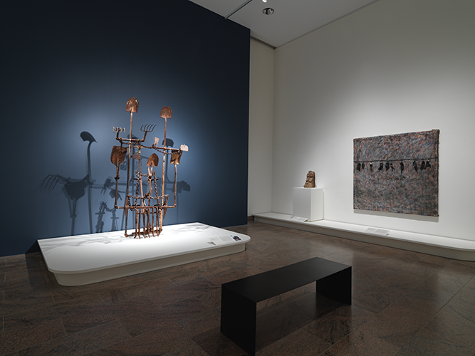 A corner view of a large metal sculpture made of work tools on a plinth with a blue wall behind it, and on the right is a small sculpture and large mixed-media artwork.