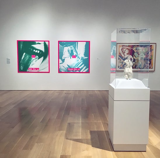 Museum gallery view of "Breaking the Mold" exhibition with paintings on the wall and a sculpture in a vitrine in the foreground.