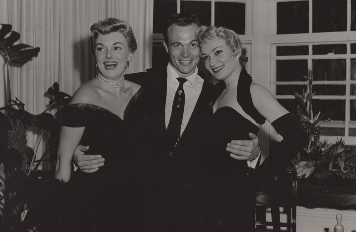 vintage Hollywood photo of a man with his arms around the waits of a women on either side of him.