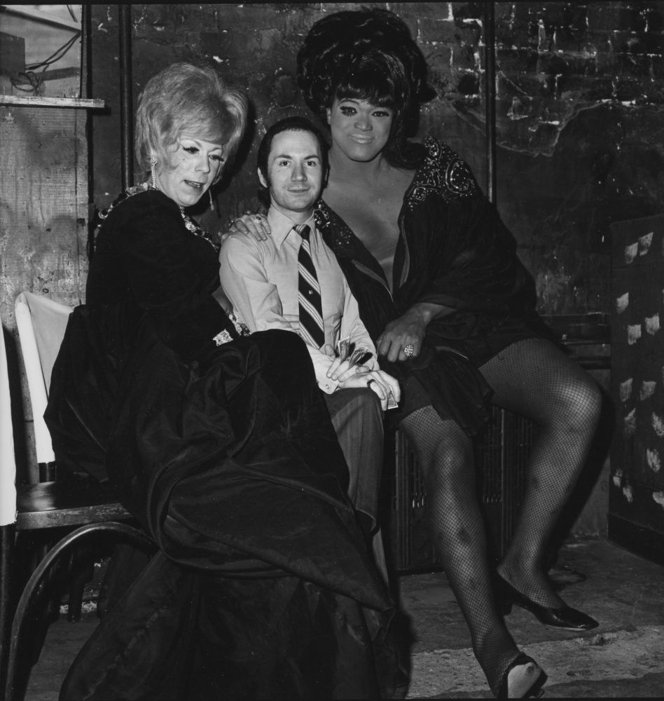 a black and white image of two drag queens embracing a man in a tie.