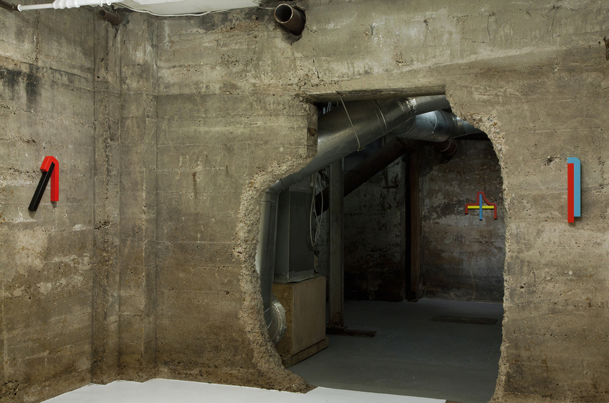 Small multicolor shaped sculptures hang in a cement gallery with strange architecture