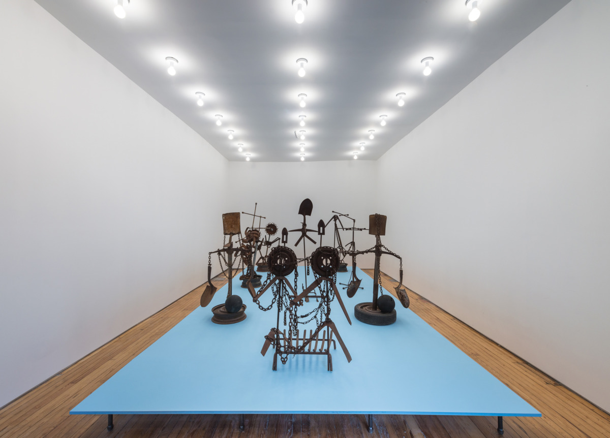 a white paneled room with a blue raised platform covered in rusted metal sculptures;
