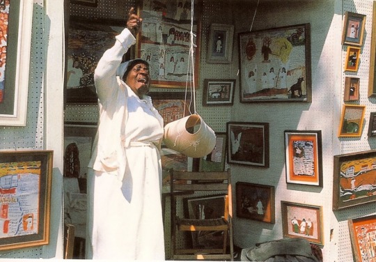 an elderly Black woman dressed in a long white skirt and long-sleeve shirt stands in front of a small room with colorful, framed paintings on the wall