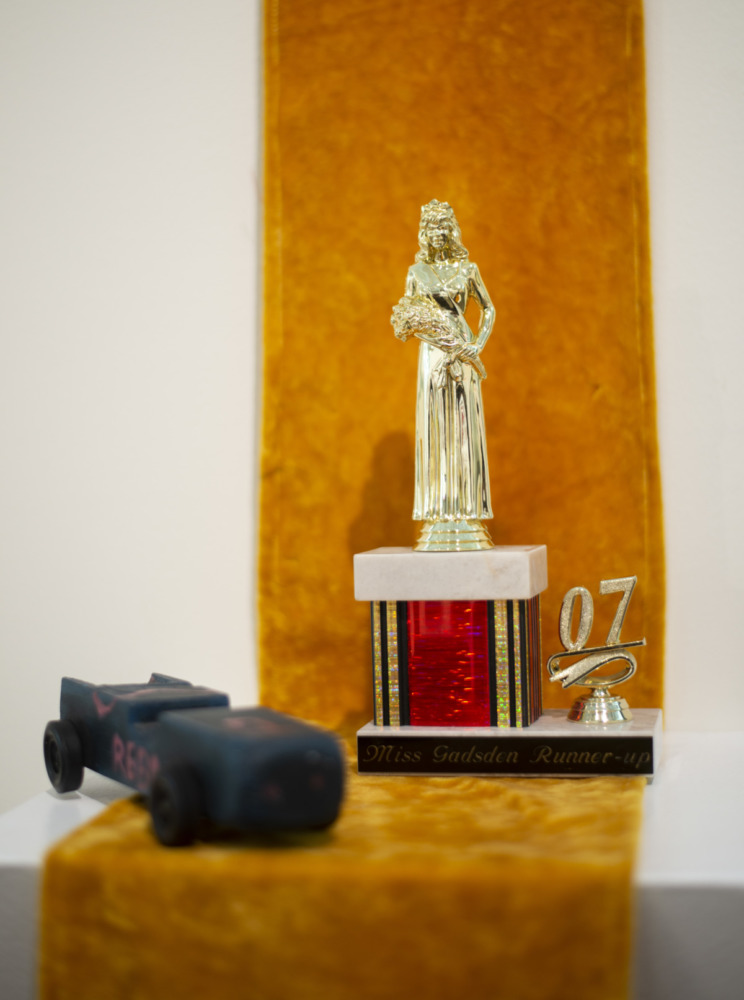 against a white wall hangs an orange velvet ribbon. on the orange velvet ribbon stands a trophy. the trophy person is a woman holding flowers. o the base of the trophy says Miss Gadsden Runner up 2007. in the foreground of the picture there is a black toy car.