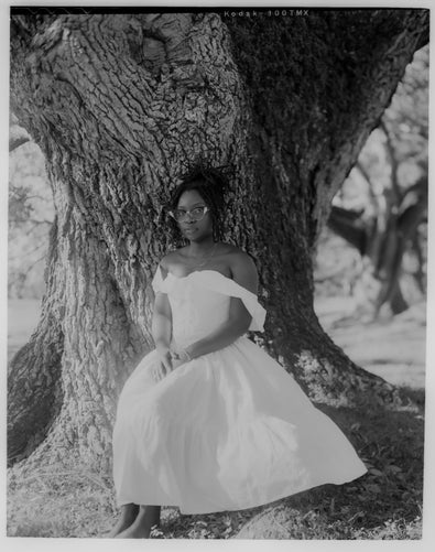 a black femme wearing ornate cat-eye glasses in a off-the-shoulder white dress against the trunk of a large tree