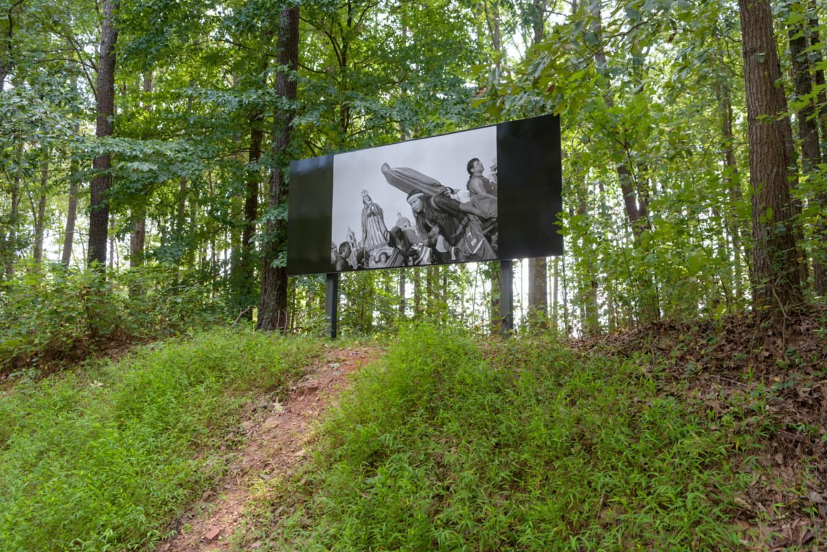 billboard installation of black and white photograph depicting a person carrying a religious statue is seen at the top of a hill surrounding a lush green forest woodland