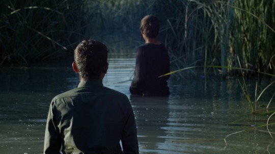 two boys waist deep in a watery marsh, turned with their clothed backs to the camera