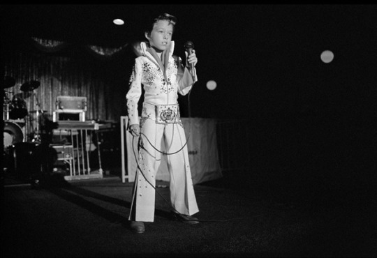 a black and white photograph of a young boy emulating Elvis on stage with wide-legged trousers and a decorated blazer
