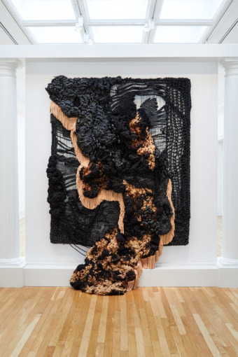 a white-walled, light-filled gallery is filled with sweeping textiles that reach from floor to ceiling, the installations are a charcoal black with a brown underlay on fabric, they appear like blooming clouds of smoke from an explosion