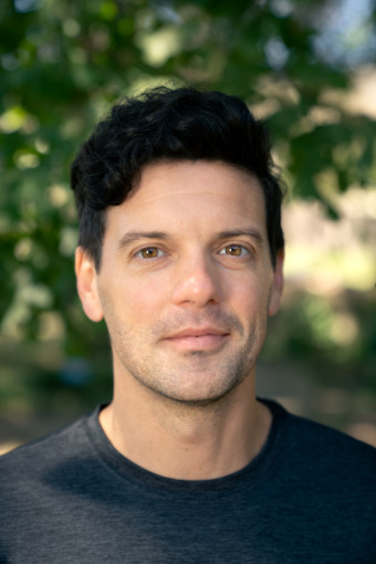 a shoulders-up headshot of Trey Burns, who wears a black shirt and stands against a slightly blurred nature background. Trey has curly black hair that is cut short and a thoughtful smile on his face
