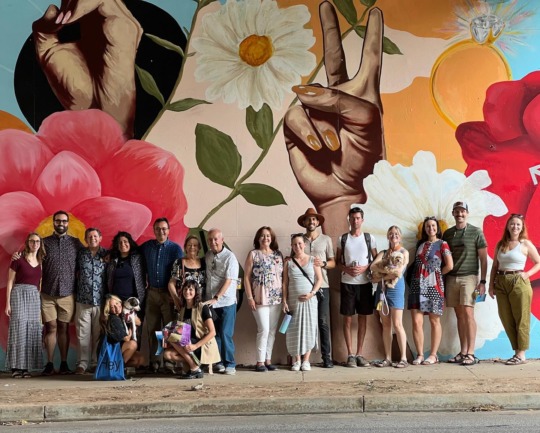 A group of friends and community members stand in front of the finished mural for the Laura Patricia Calle Grant. The details behind them include flowers and hands spelling out “love” in ASL.