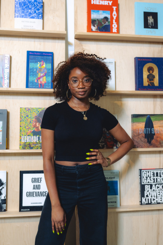 a Black woman wears a cropped black t-shirt and dark pants with a necklace. her bright manicured hand rests on her hip. she stares at the camera with a thoughtful gaze with wide-rimmed glasses and a curly brown fro