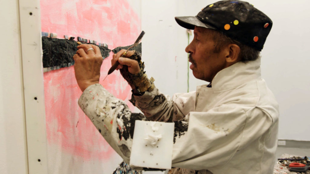 black male artist chisels away at tessarae on a pink canvas