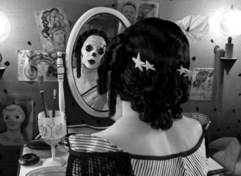 black and white image of woman dressed with button eyes and staring at her reflection in a vanity mirror